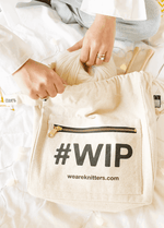 The #WIP Bag included in this bundle - cream cotton canvas project bag with a front zip pocket, a ball of yarn being lowered inside
